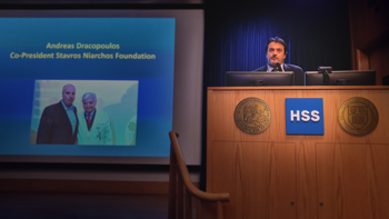 Ένας άντρας μιλάει στο βήμα του Hospital for Special Surgery και δείχνει στον προτζέκτορα τη φωτογραφία του Ανδρέα Δρακόπουλου, Πρόεδρο του Ιδρύματος Σταύρος Νιάρχος μαζί με τον τιμώμενο καθηγητή Παναγιώτη Κουλουβάρη